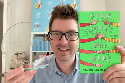 A man in a blue shirt and glasses holds up a trophy and book titled, Finn Jones Was Here, and smiles